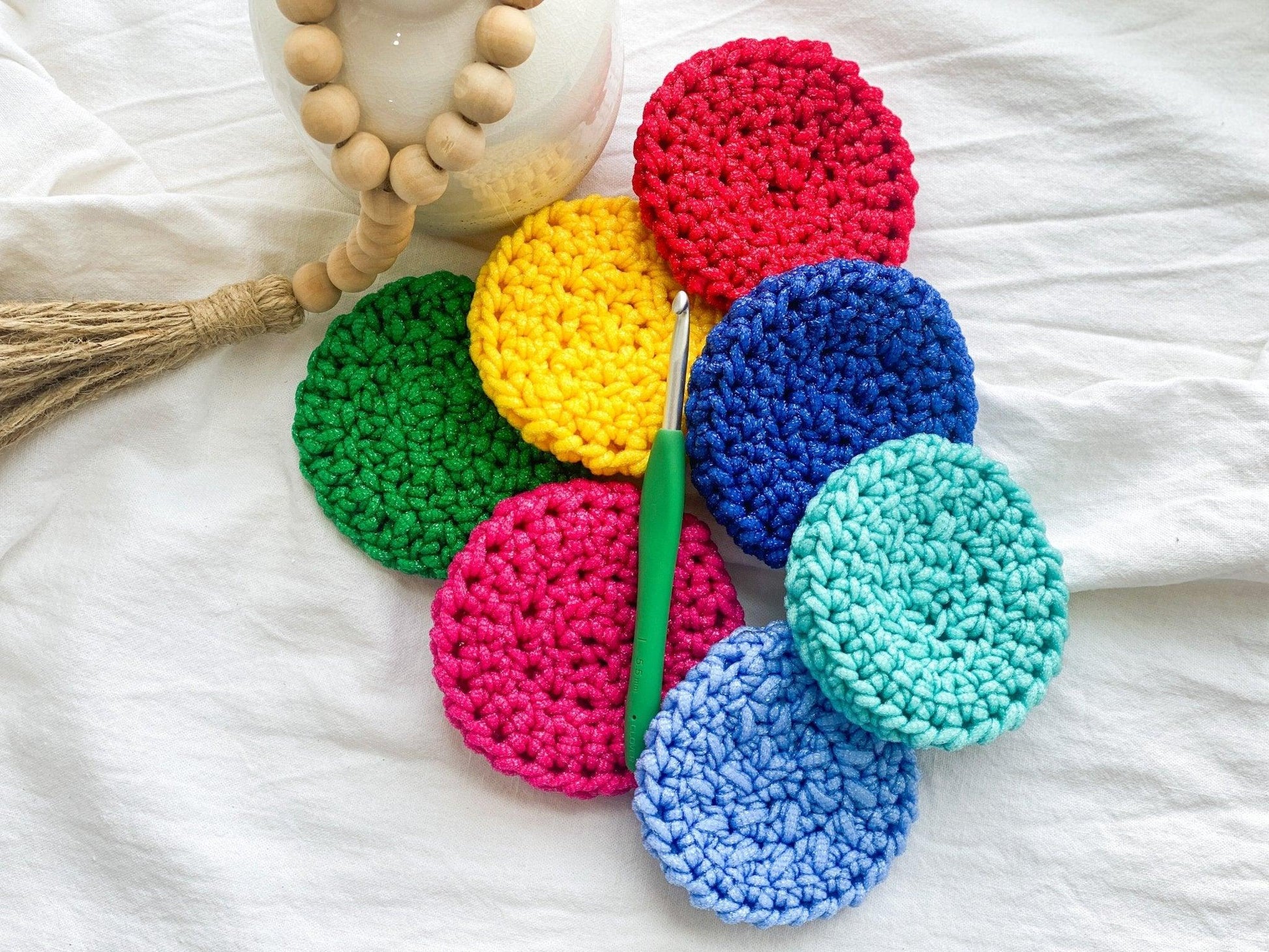 Several Bright Handmade Crochet Dish Scrubbies laying on White Linen Cloth with Green Handled Crochet Hook