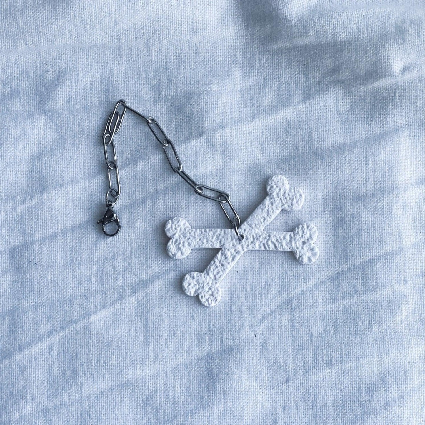 White Crossbones Stanley Cup Charm Attached To Silver Stainless Steel Chain Laying Flat on White Linen Cloth