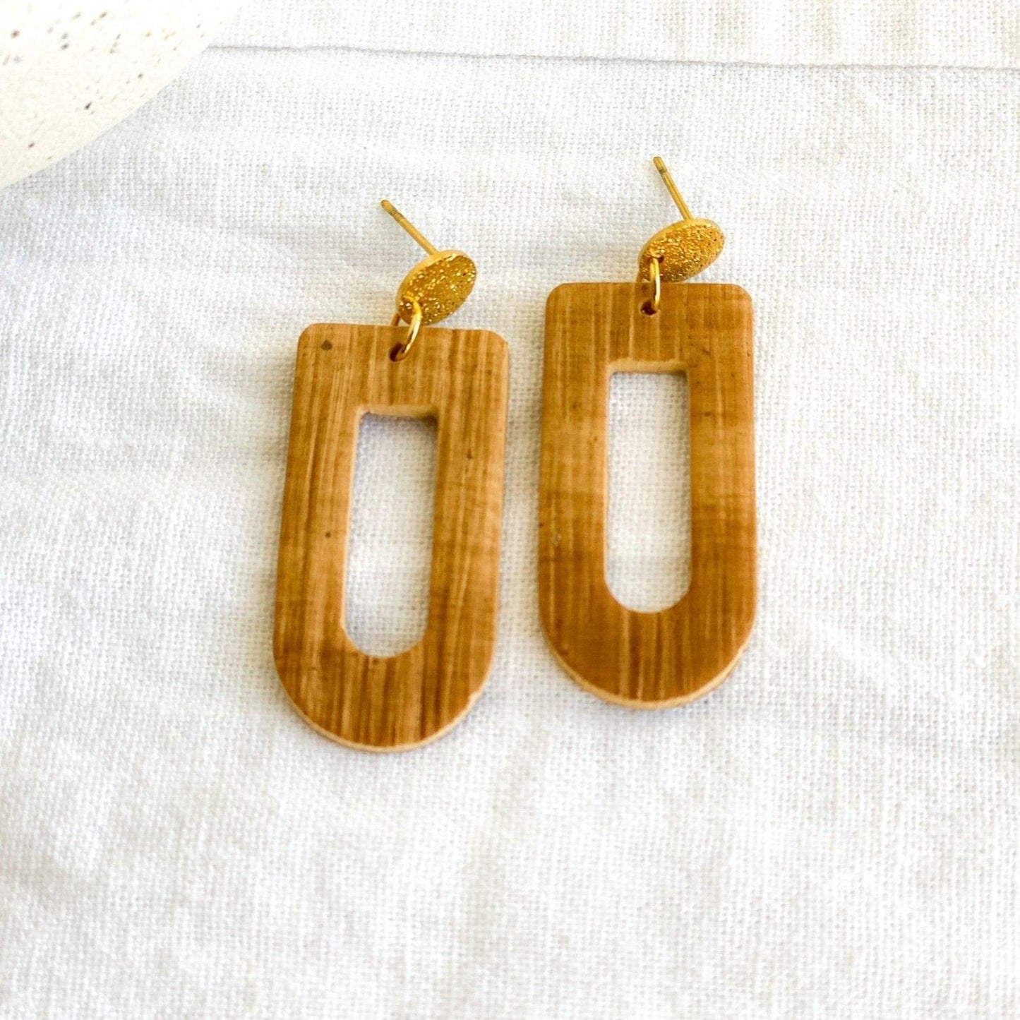 close up of cute handmade brown earrings with gold surgical steel posts laying on white cloth next to white vase