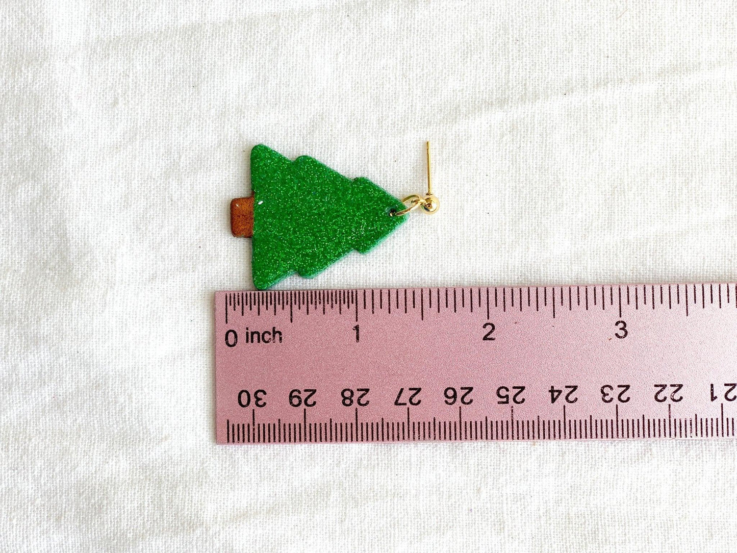 handmade earrings that are sparkly green christmas trees attached to gold ball posts next to pink ruler on white cloth