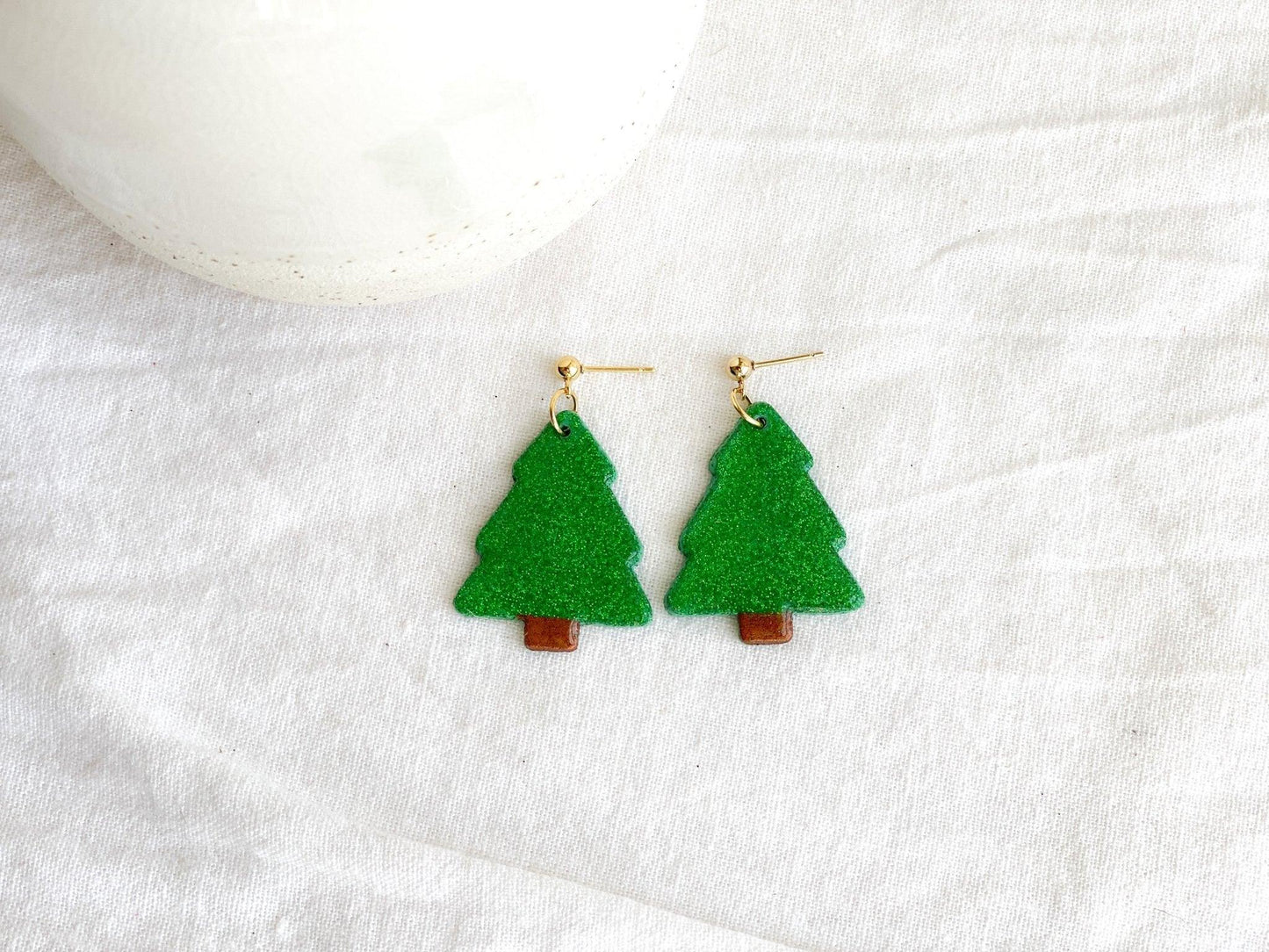 handmade earrings that are sparkly green christmas trees attached to gold ball posts on earring card on white linen cloth