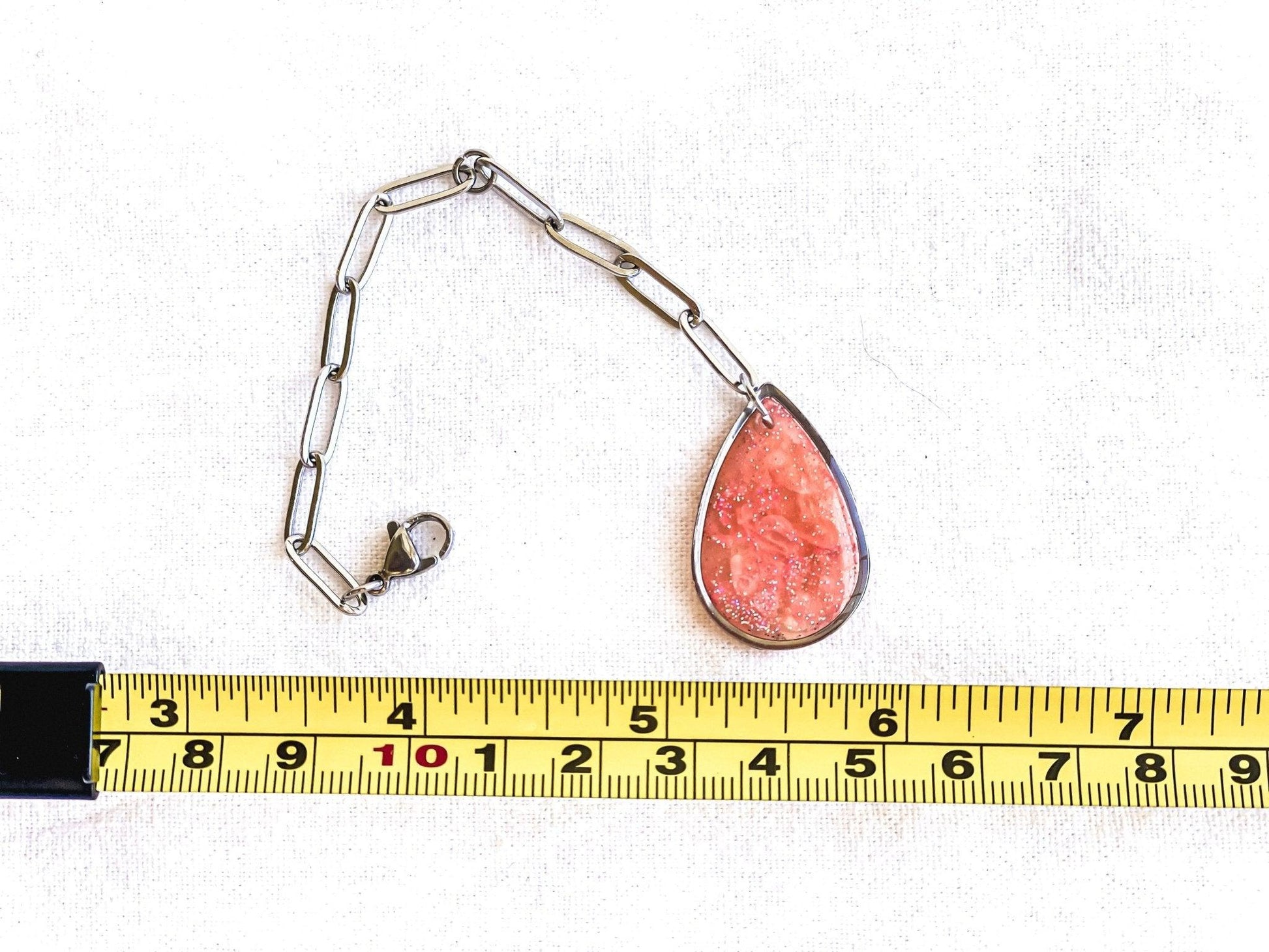 Pink Teardrop Stanley Cup Charm Attached To Silver Stainless Steel Chain Laying Flat On White Cloth Next To Measurement Tape