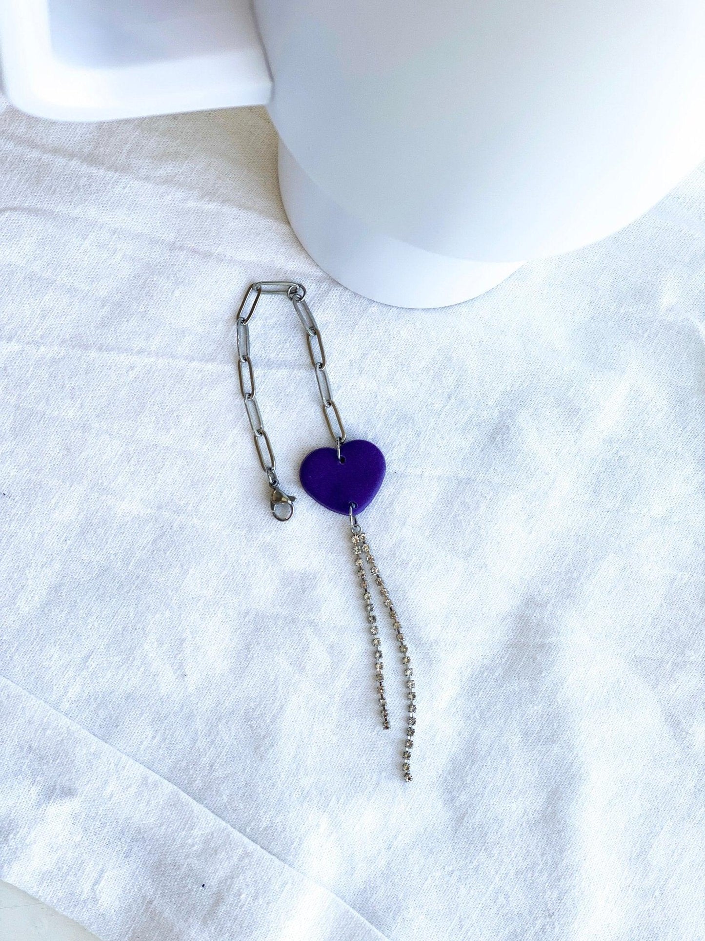Purple Heart with Silver Rhinestone Strands Stanley Charm Attached To Silver Stainless Steel Chain Laying Flat on White Cloth