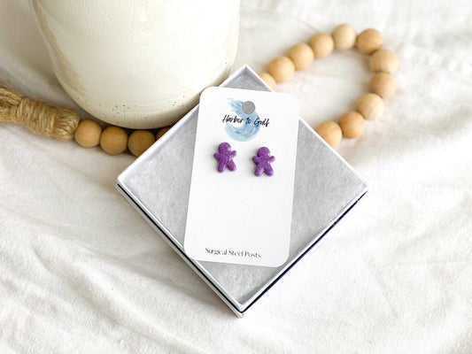 small sparkly purple handmade gingerbread men stud earrings on earring card in white gift box propped on strand of wood beads