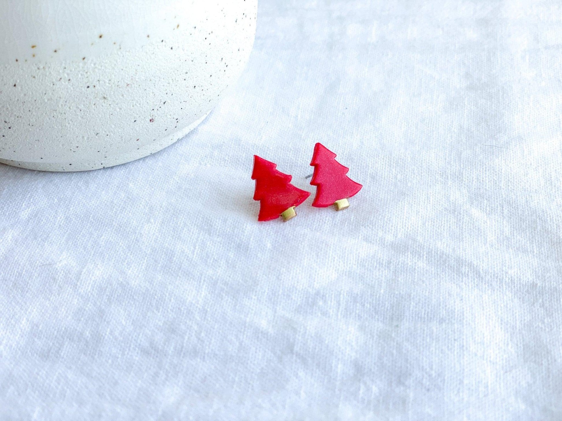 Red Stud Earrngs shaped like Christmas Trees with surgical steel posts on white cloth next to white vase