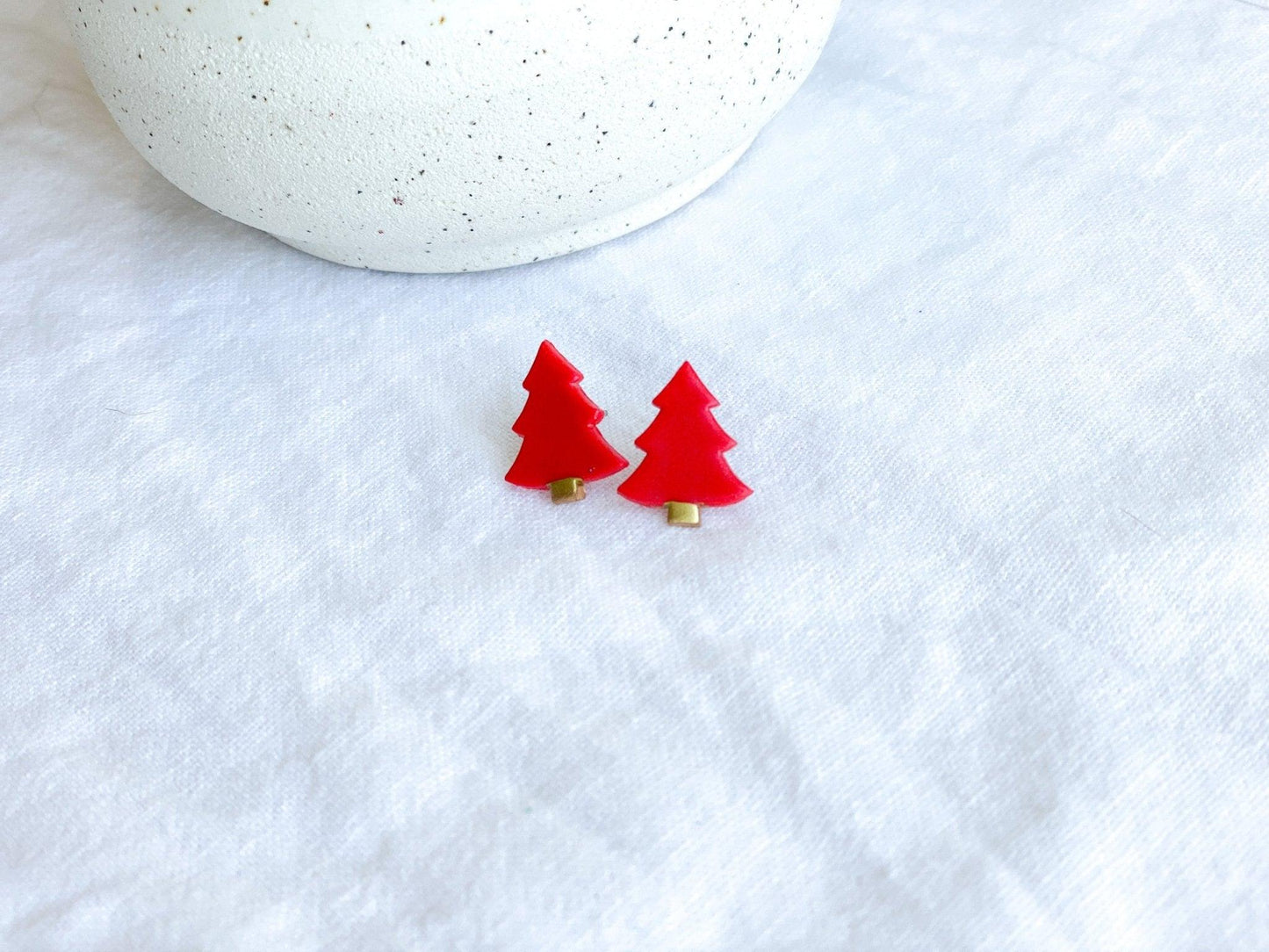 Red Stud Earrngs shaped like Christmas Trees with surgical steel posts on white cloth next to white vase