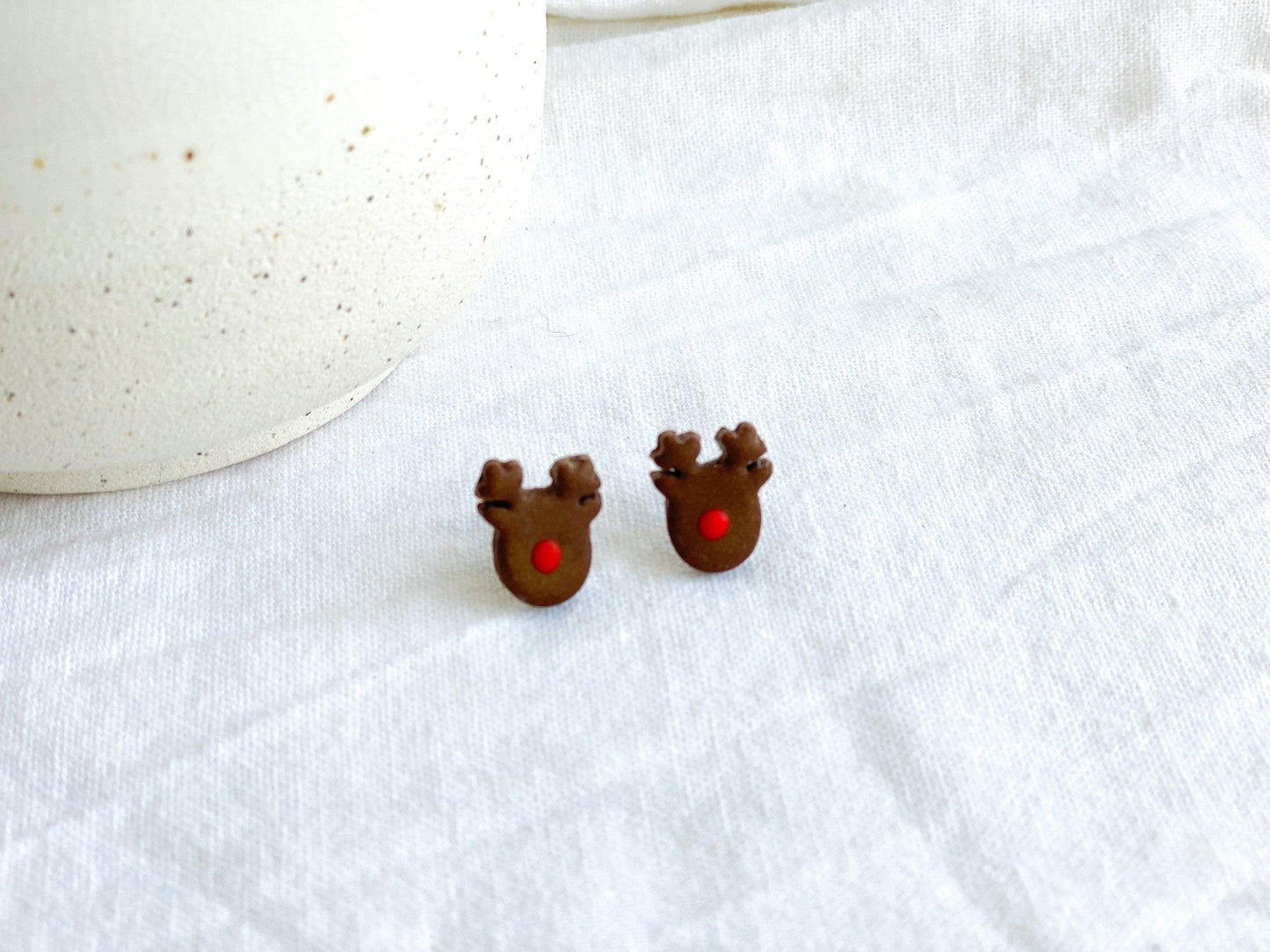 Cute handmade Rudolph Stud Earrings made from polymer clay and surgical steel posts on white cloth next to white vase