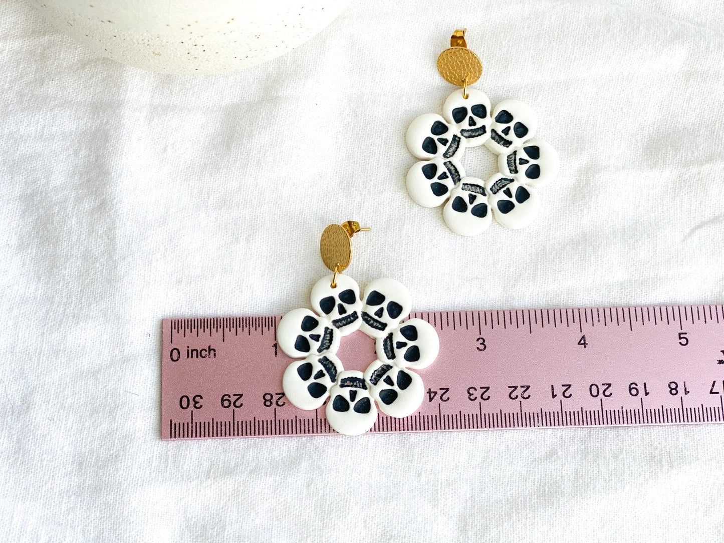 Handmade White and Black Skull Circle Earrings with Gold Surgical Steel Circle Stud Tops on White Cloth Next to Pink Ruler