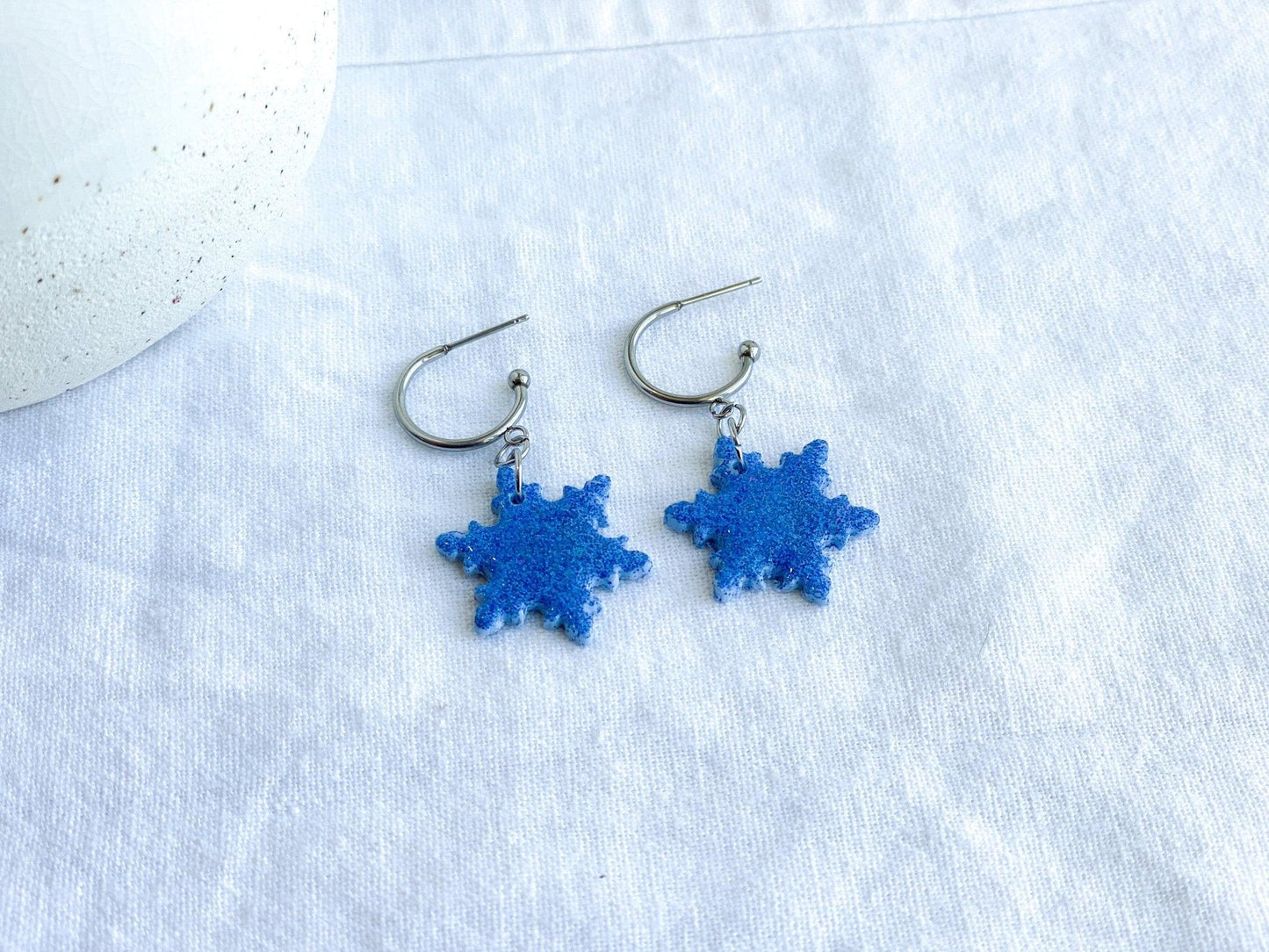 Handmade Sparkly Blue Snowflake Earrings on Silver Surgical Steel Huggie Earrings on White Linen Cloth