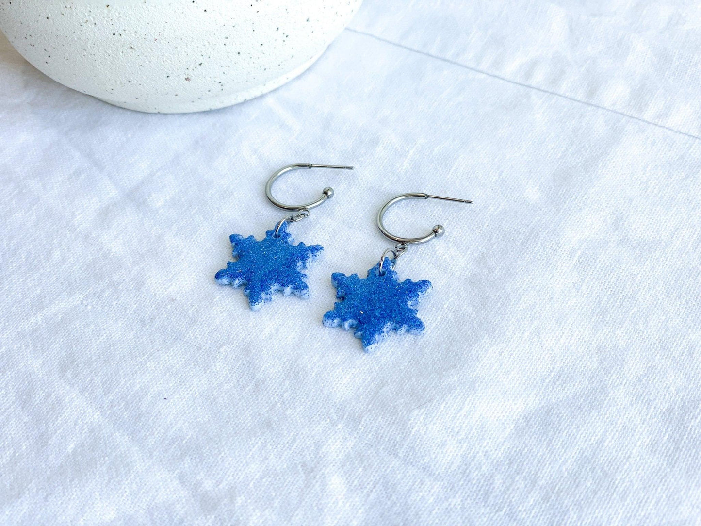 Handmade Sparkly Blue Snowflake Earrings on Silver Surgical Steel Huggie Earrings on White Linen Cloth