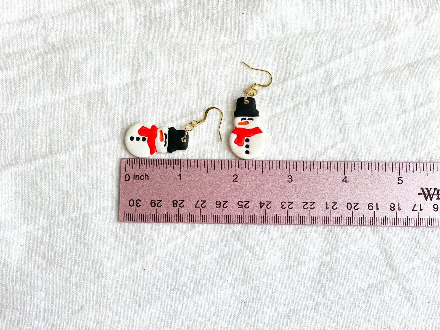 Cute Handmade Clay Snowman Earrings on Gold Ear Wires made from Surgical Steel laying on white cloth next to pink ruler