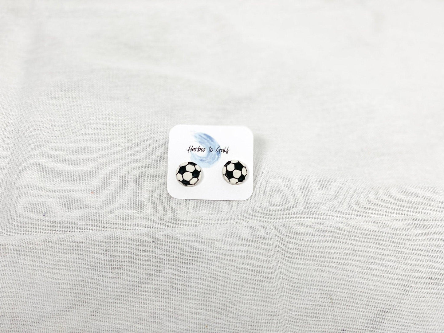 Small, Handmade Soccer Stud Earrings with Surgical Steel Posts on Harbor to Gulf Earring Card laying on White Linen Cloth