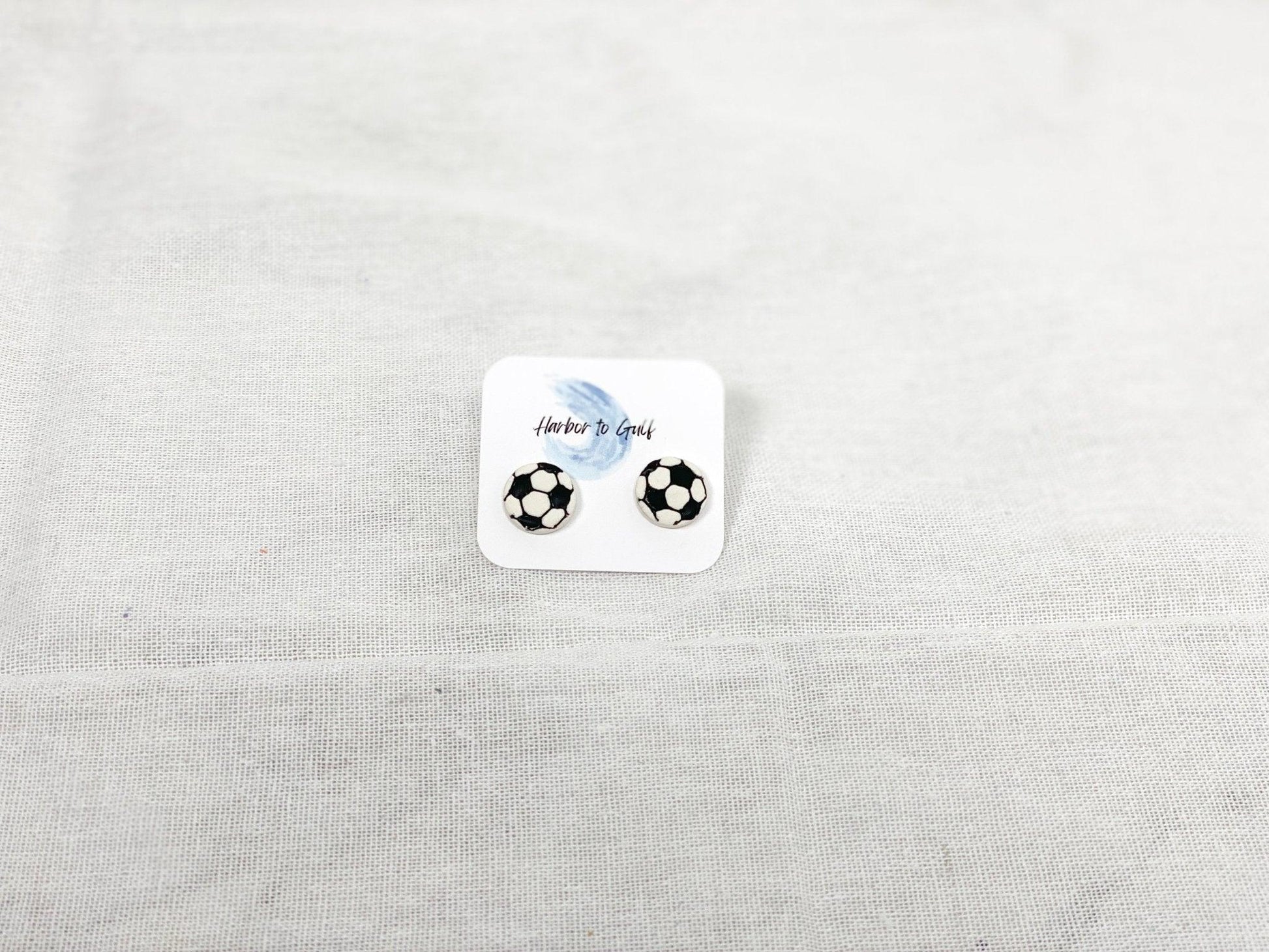 Small, Handmade Soccer Stud Earrings with Surgical Steel Posts on Harbor to Gulf Earring Card laying on White Linen Cloth