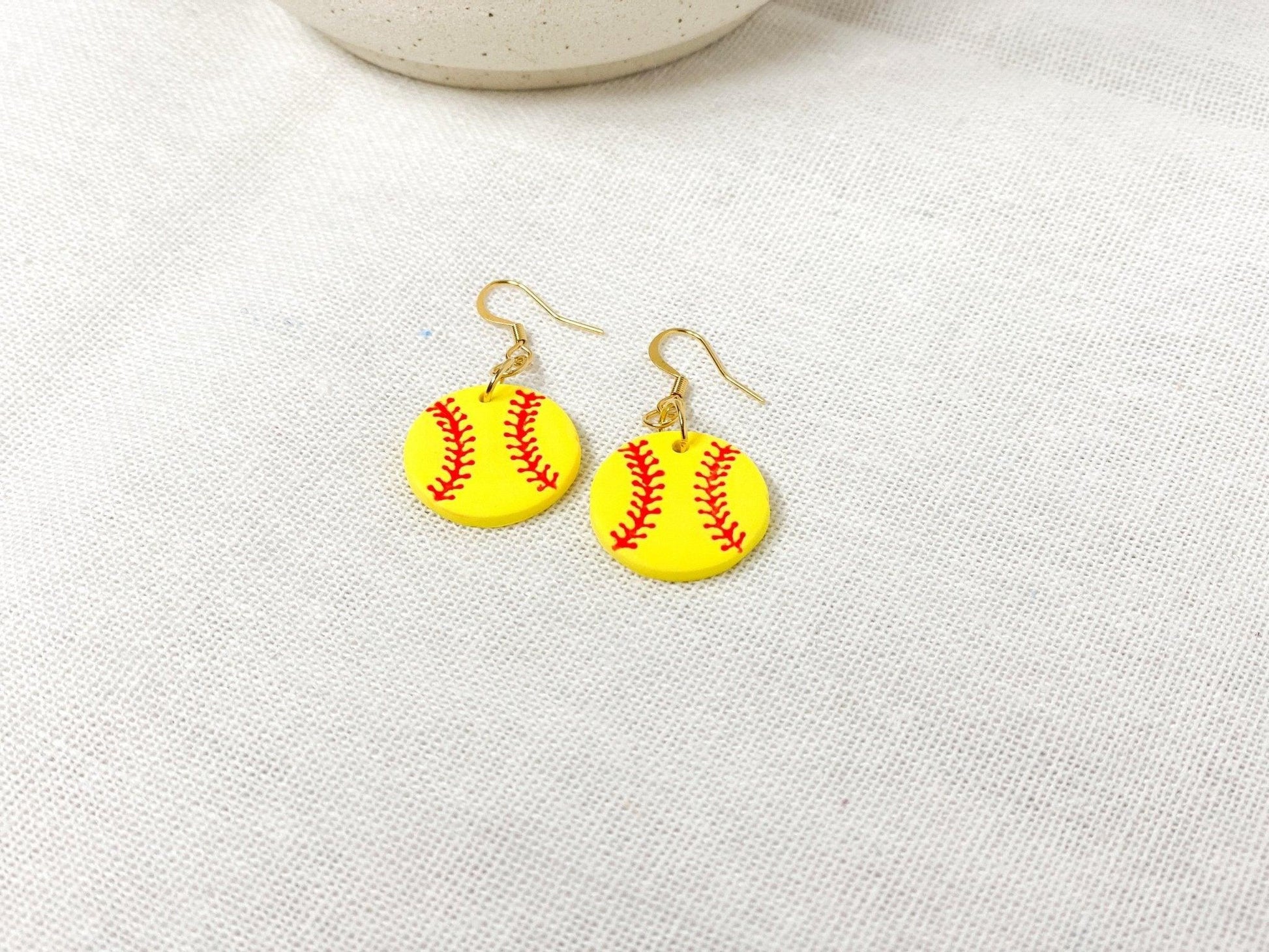 Handmade Softball Earrings with Gold Surgical Steel Ear Wires laying flat on White Linen Cloth