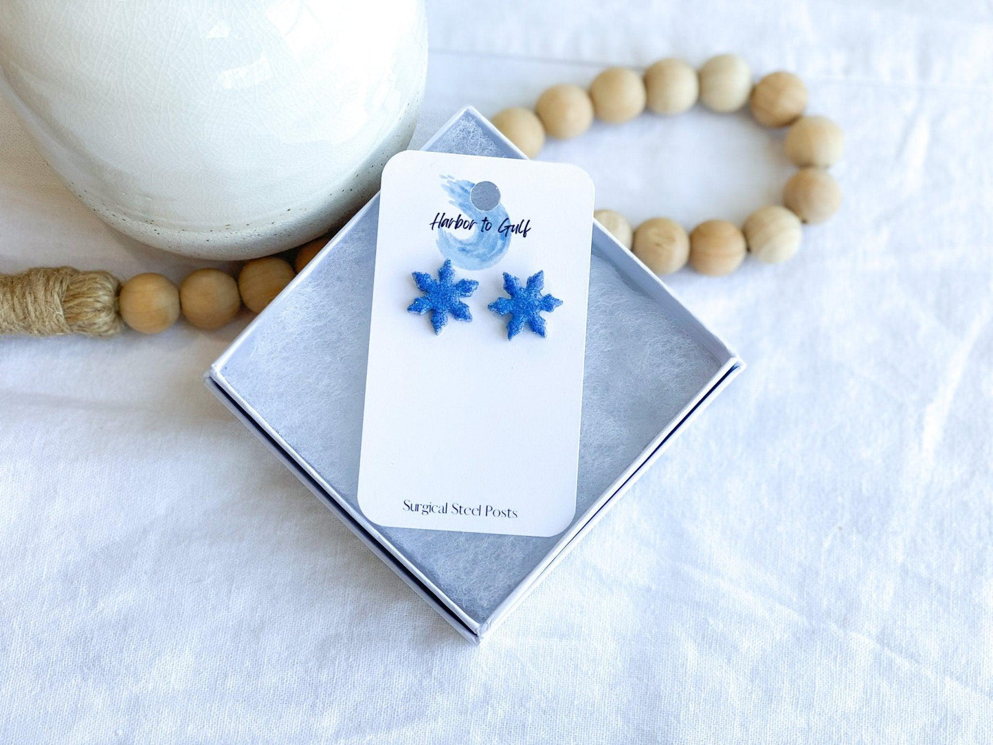 small handmade blue sparkly snowflake stud earrings in on earring card that states surgical steel studs in white gift box