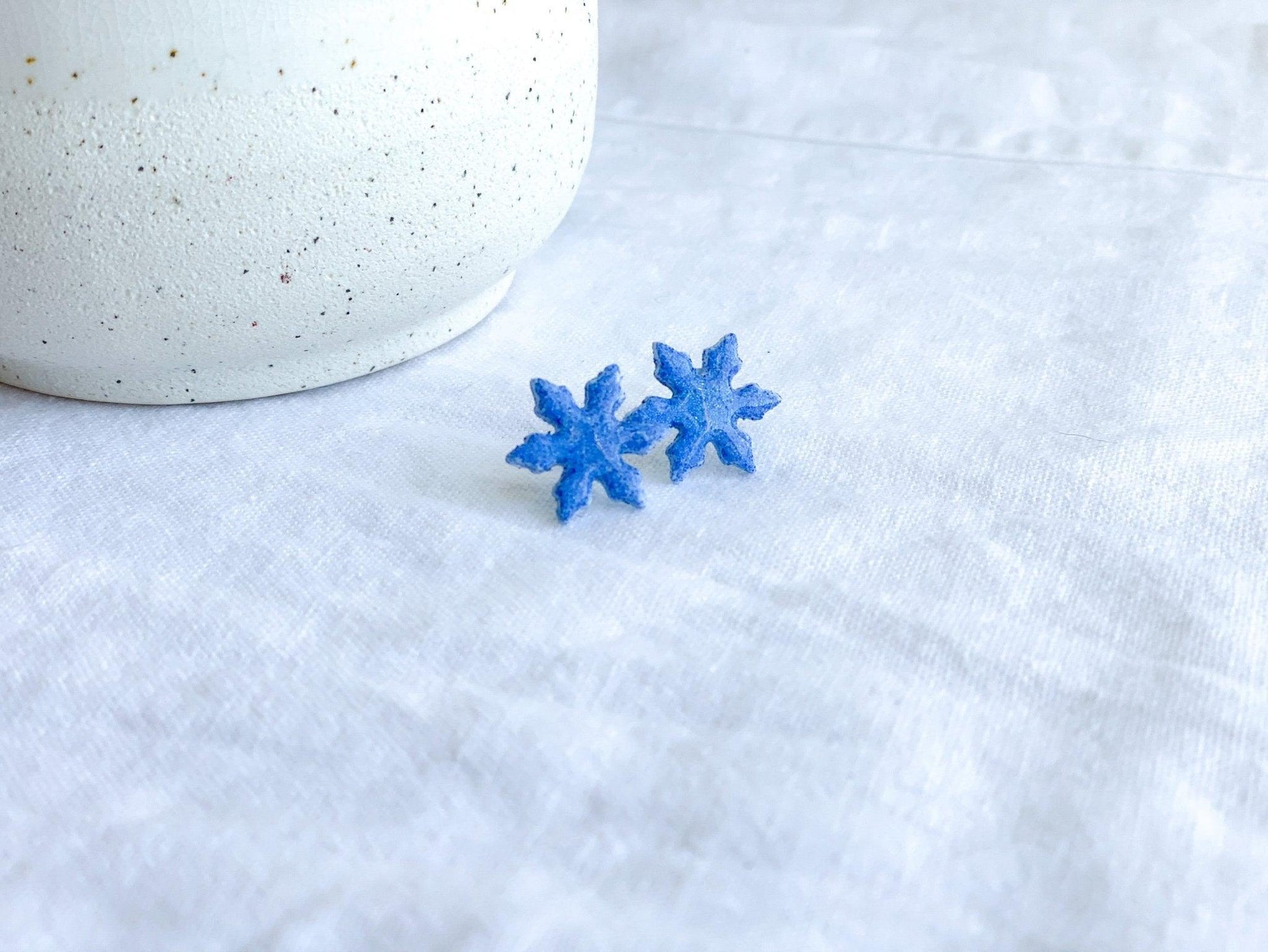 small handmade blue sparkly snowflake stud earrings on earring card that states surgical steel studs on white linen cloth