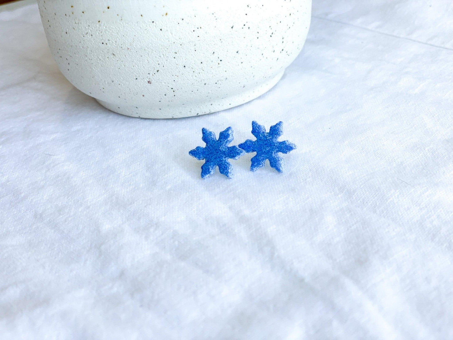 small handmade sparkly snowflake stud earrings on earring card that states surgical steel studs on white linen cloth