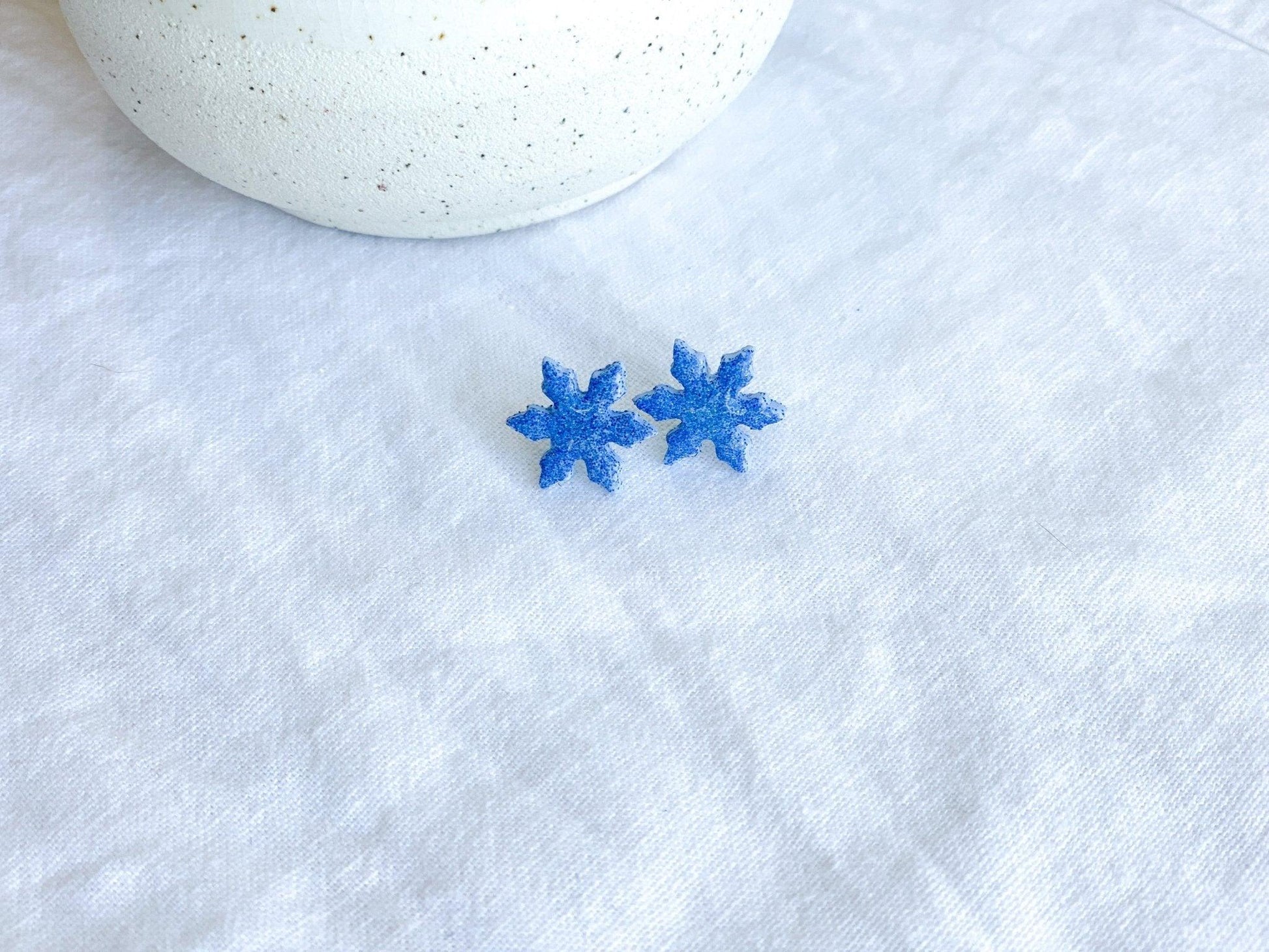 small handmade sparkly snowflake stud earrings on earring card that states surgical steel studs on white linen cloth