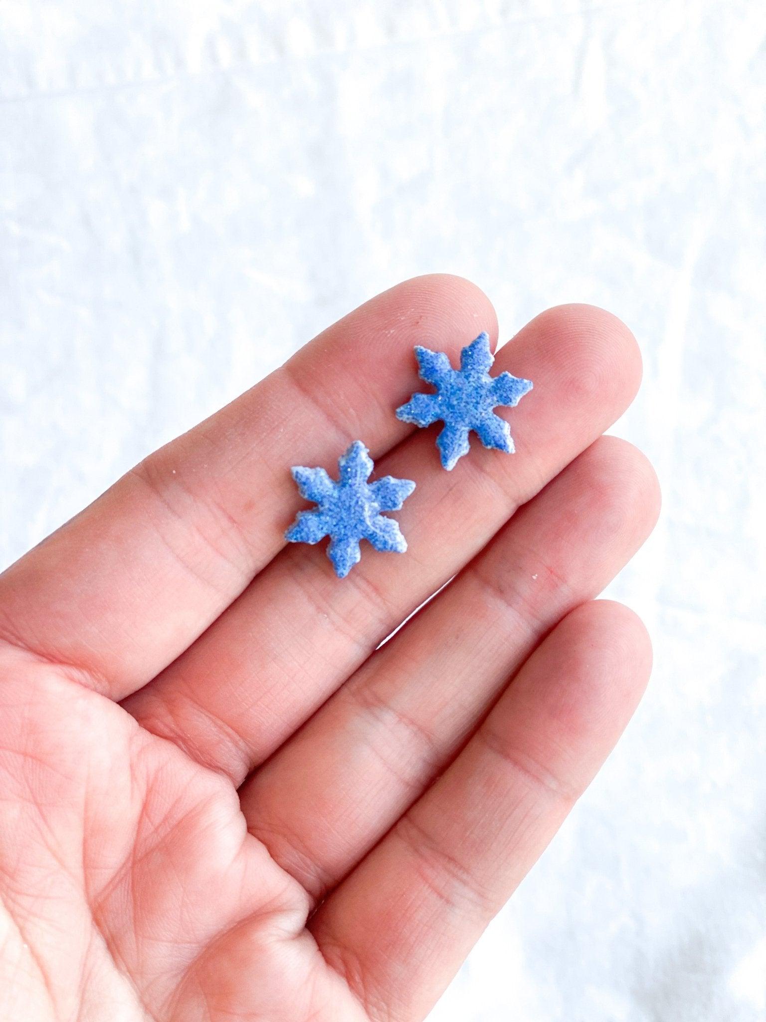 small handmade sparkly snowflake stud earrings on earring card that states surgical steel studs on human hand for size