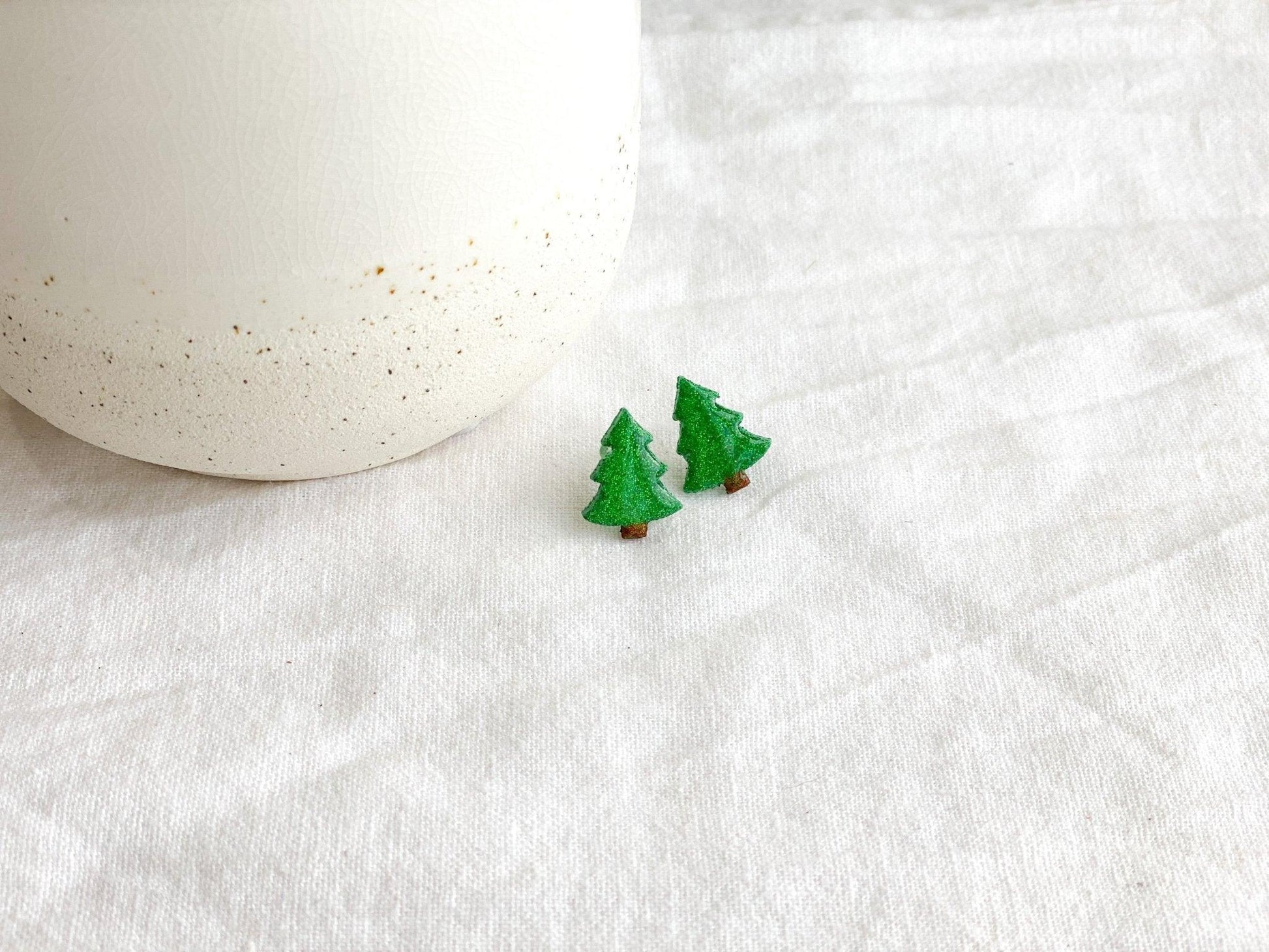 Small Handmade Green Sparkly Christmas Tree Stud Earrings on White Linen Cloth next to white ceramic jar