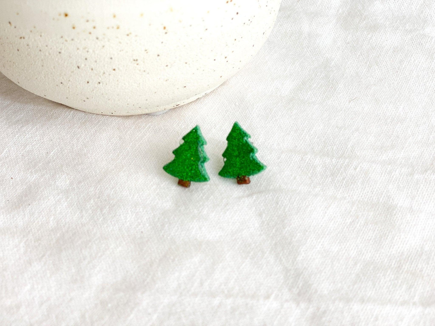 Small Handmade Green Sparkly Christmas Tree Stud Earrings on White Linen Cloth next to white ceramic jar