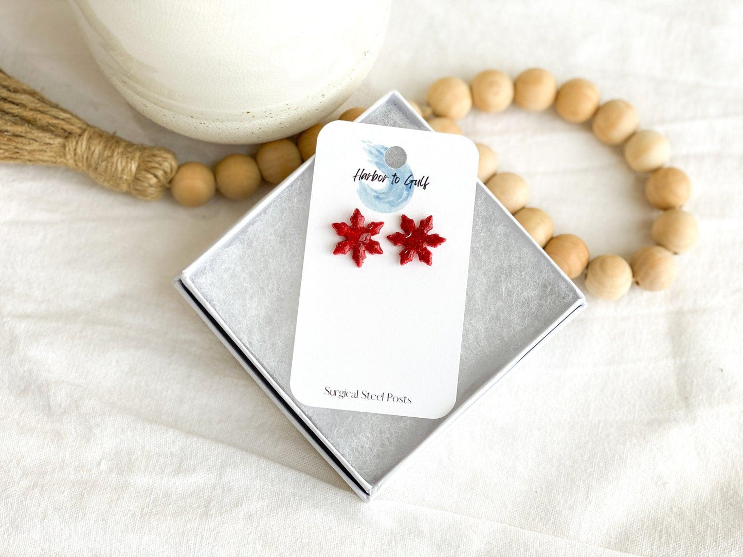 Small Handmade Red Sparkly Snowflake Stud Earrings on wihte earring card in white gift box propped on strand of wood beads