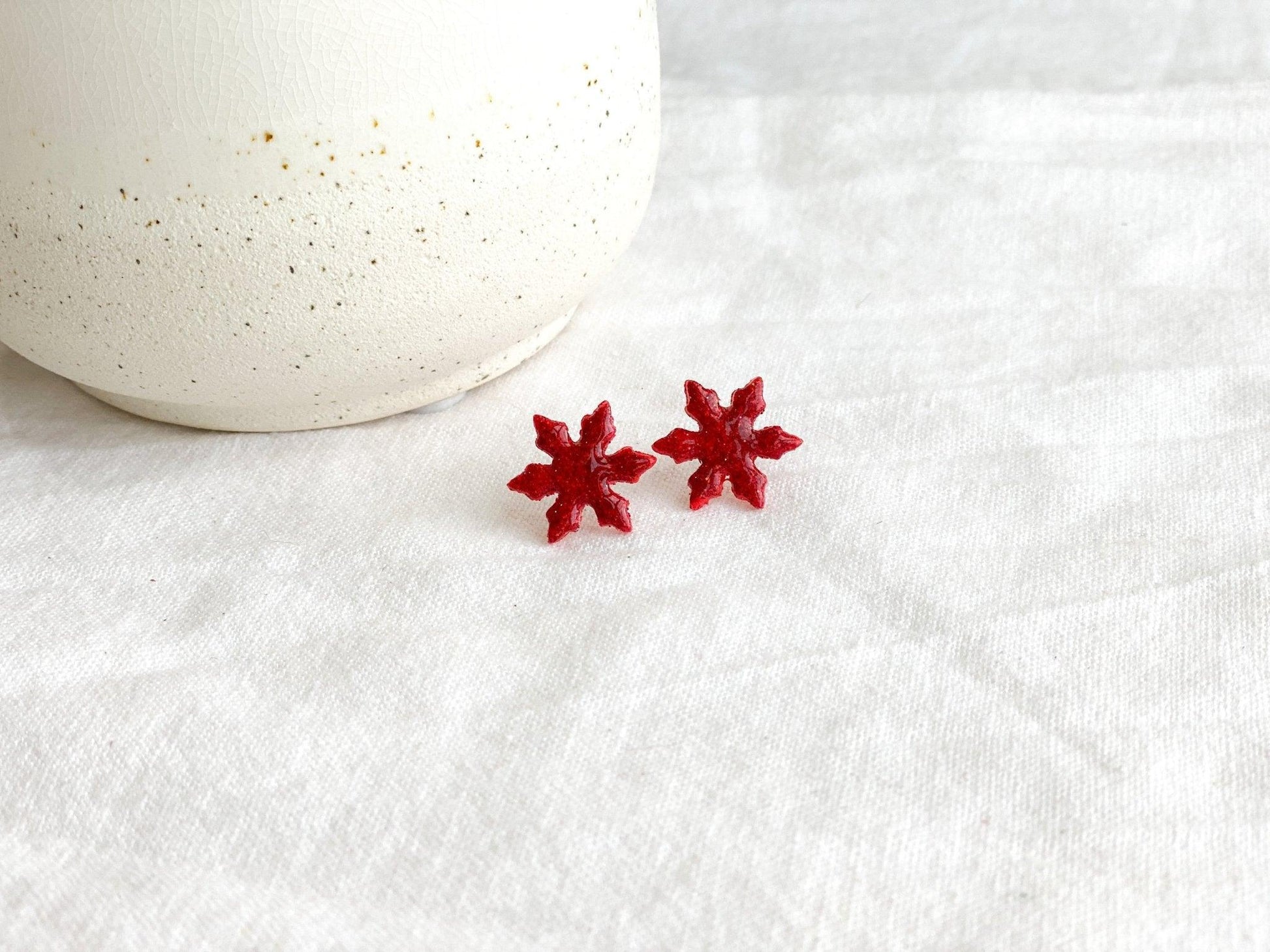 Small Handmade Red Sparkly Snowflake Stud Earrings on wihte linen cloth next to white ceramic jar
