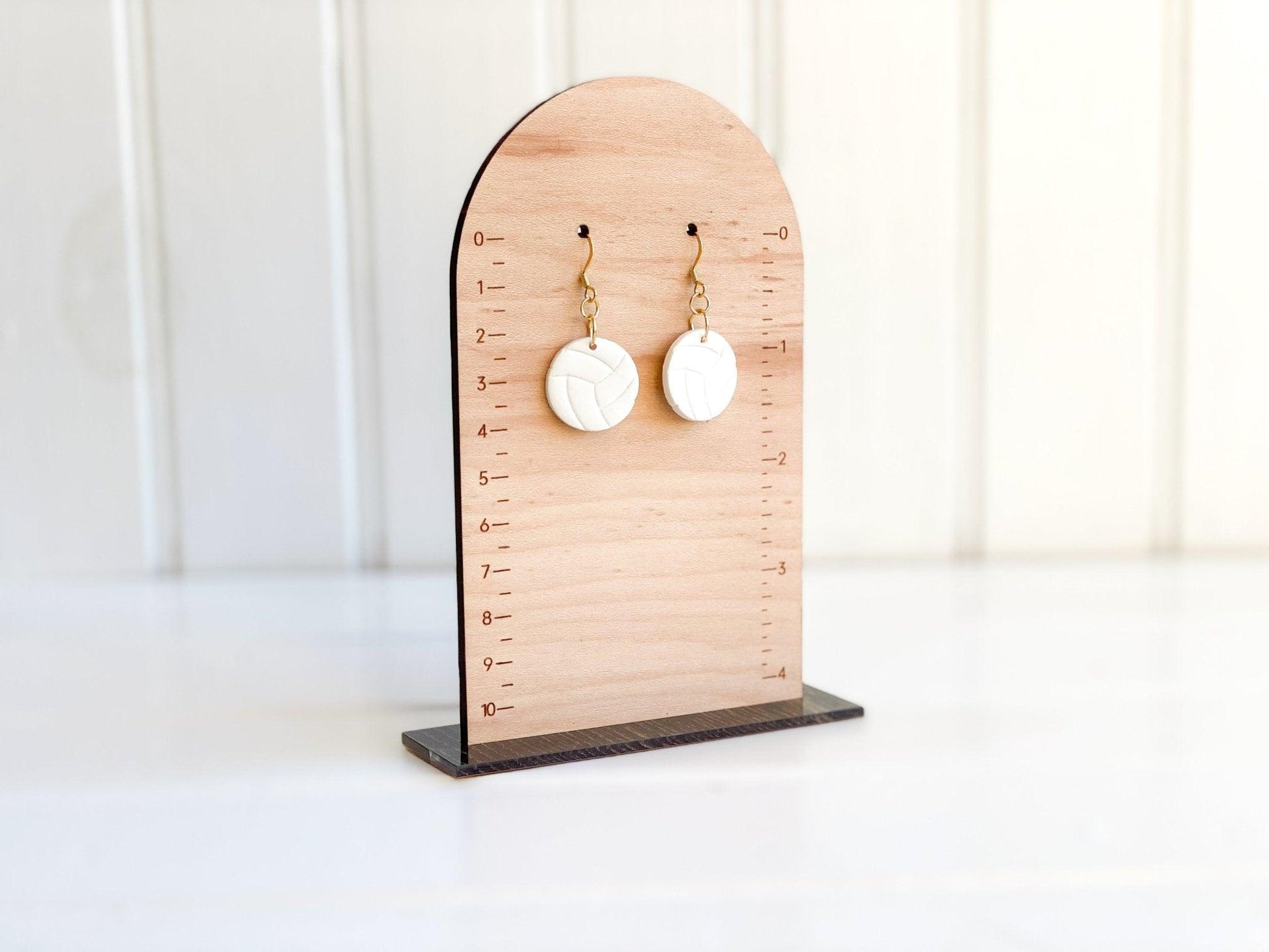 Handmade White Volleyball Earrings with Gold Surgical Steel Ear Wires Laying Flat on Wood Earring Measurement Stand