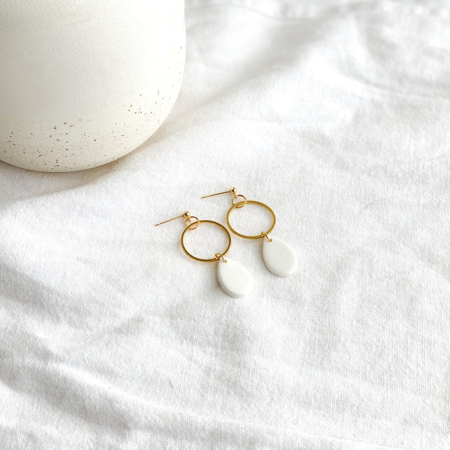 handmade white tear drops attached to thin open gold circles on gold ball posts laying flat on white cloth next to white vase